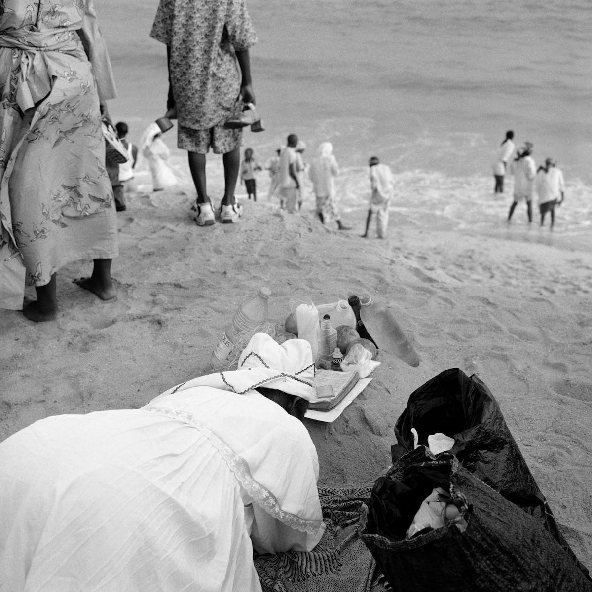 Square grainy frame on a beach: a woman in a white church robe and lace-edged headwrap bowed fully face-down in prostrate prayer on the sand, in front of an improvised altar of plastic water bottles, oil bottles and an open book; other women's legs cut the top edge, white-robed worshippers and bathers scattered in the surf behind